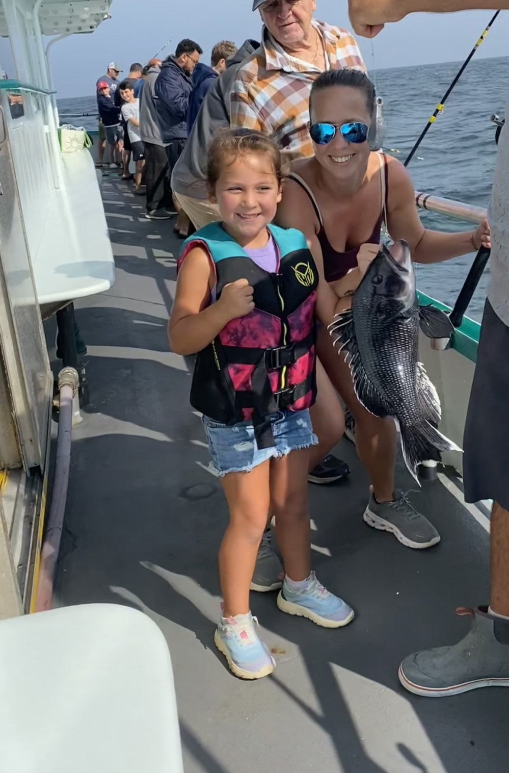 Child in life vest smiles with woman on a boat holding a large fish, surrounded by people fishing.