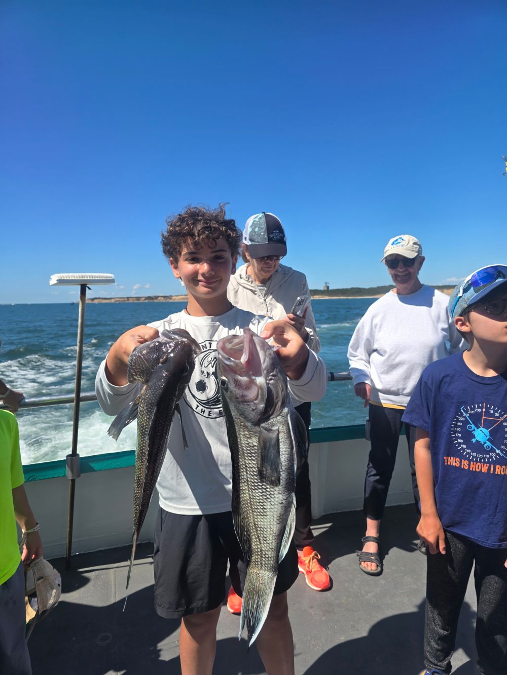 Boy on boat holding two large fish, with other people in background under a clear blue sky.
