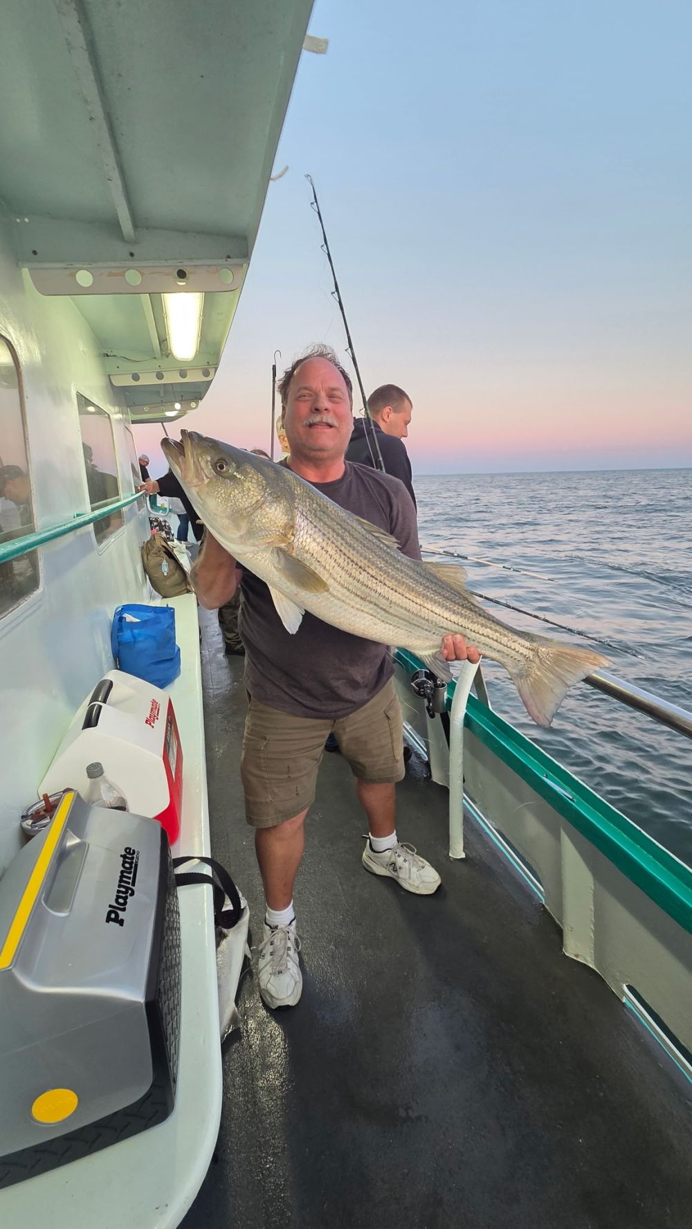 Man on boat holding large fish with ocean in background during sunset.