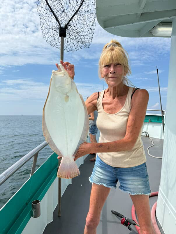 Person on boat holding a large flatfish beside a net under a blue sky.