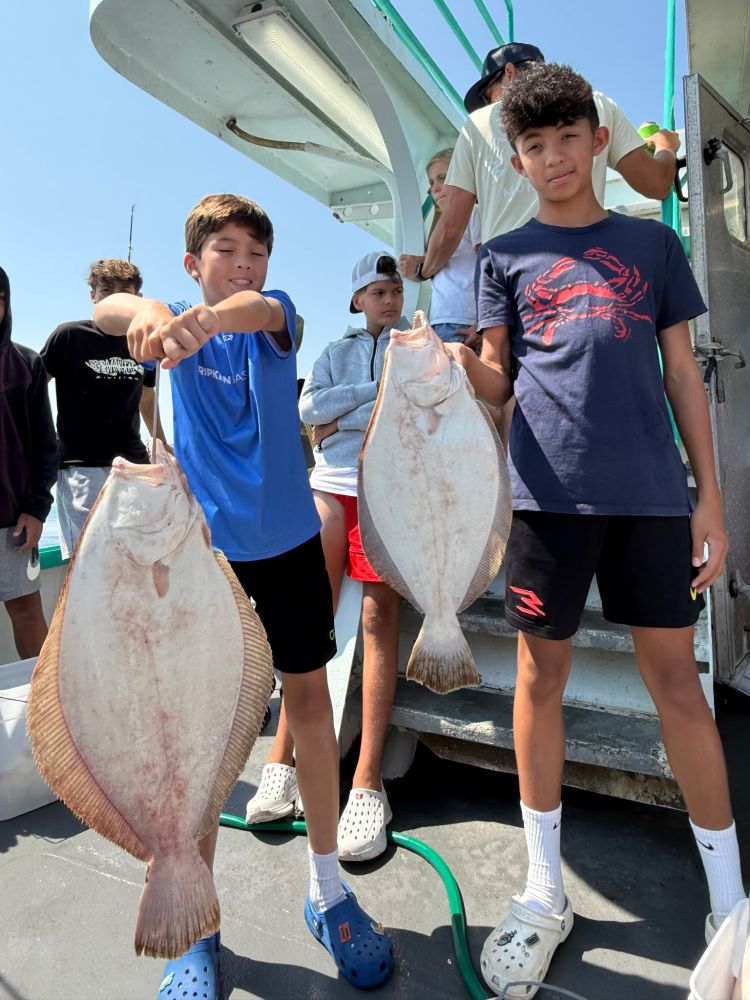 Two boys on a boat holding large flatfish; others in the background.