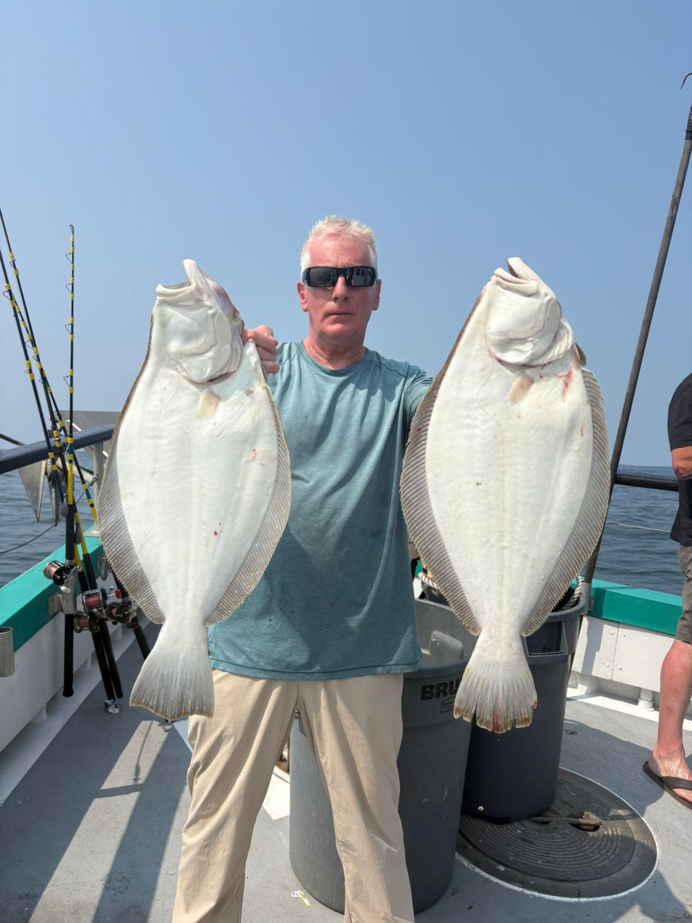 Person holding two large fish on a boat with fishing rods in the background.