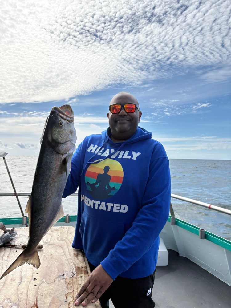 Person on boat holding a large fish under a cloudy sky, wearing a blue hoodie and sunglasses.