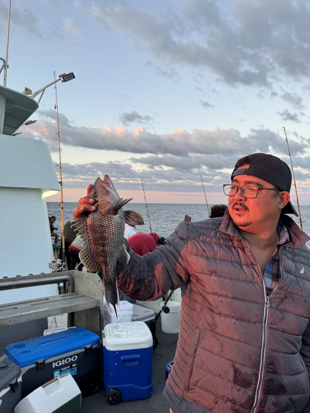 Person in jacket holding a fish on a boat at sunset, with coolers and fishing rods in the background.