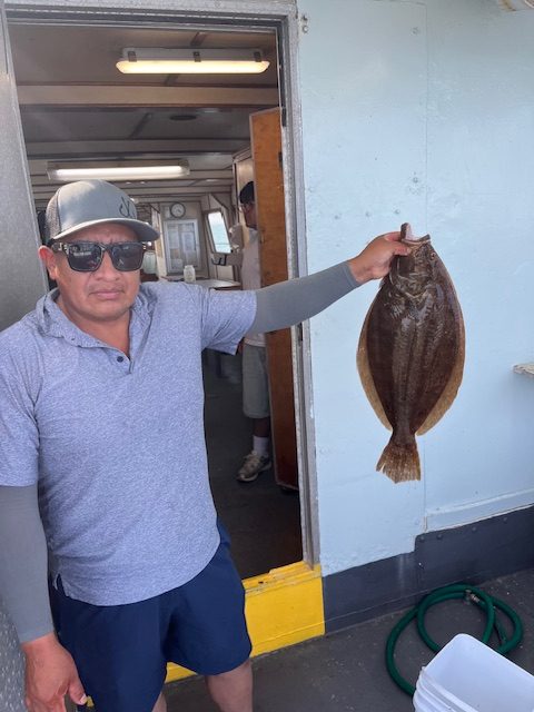 Person holding a flat fish on a boat near an open door, wearing sunglasses and a cap.