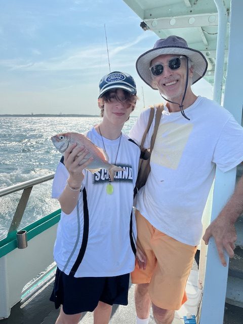 Two people on a boat, one holding a fish, both smiling under bright sunlight.