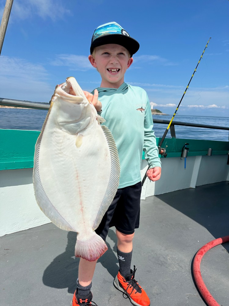 Smiling boy holding a large flat fish on a boat with a fishing rod under a clear blue sky.