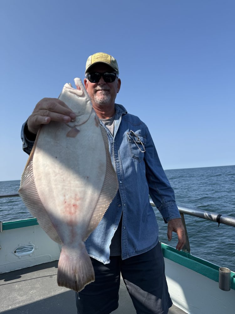 Man on boat holding a large flatfish under clear blue sky.