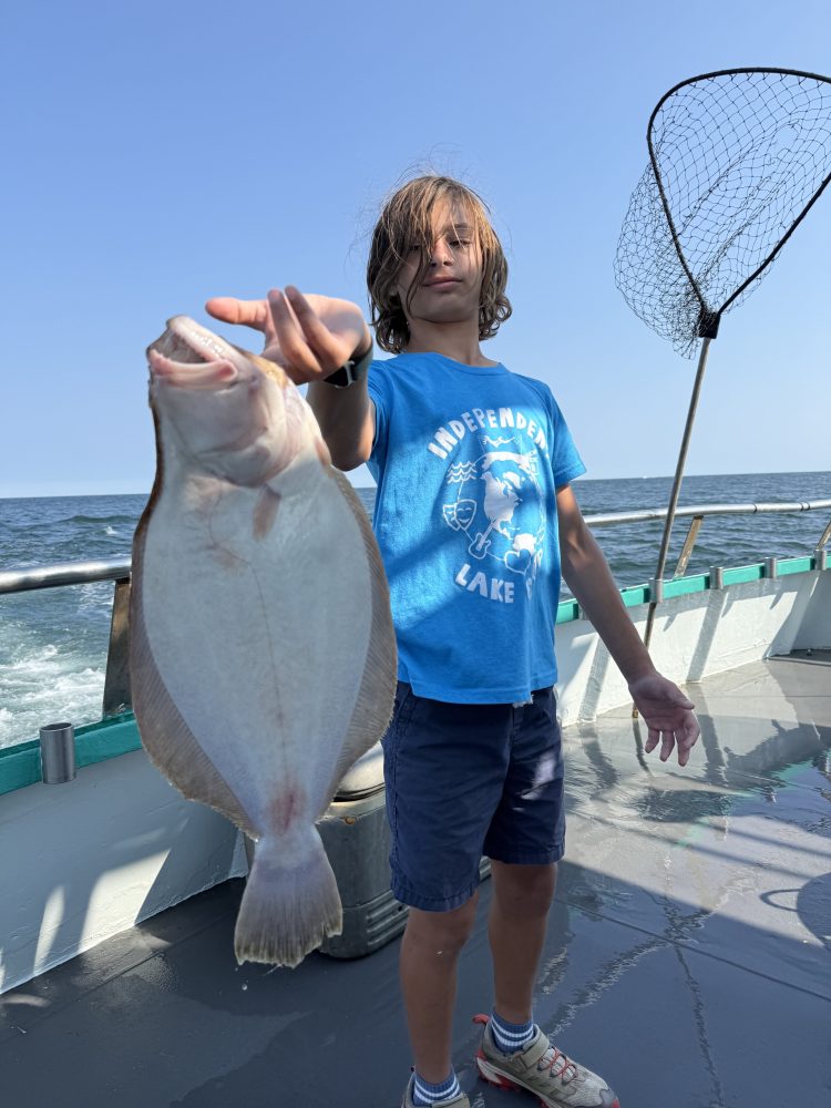 Person on a boat holding a flat fish with a net in the background under a clear blue sky.