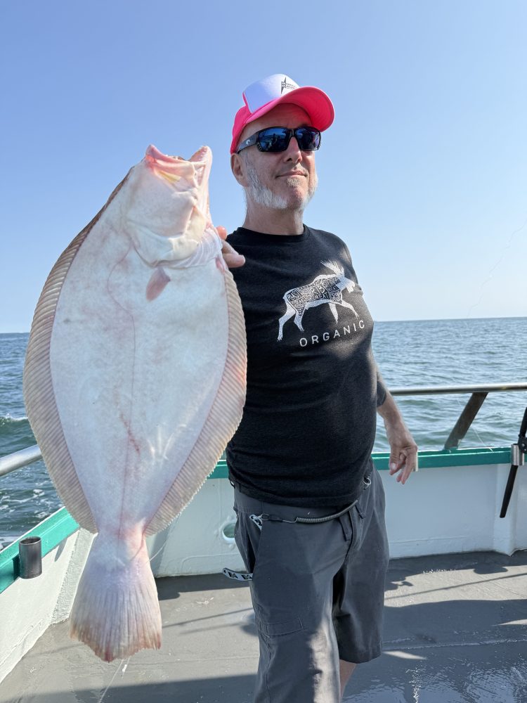 Man on a boat holding a large fish, wearing sunglasses and a pink cap.