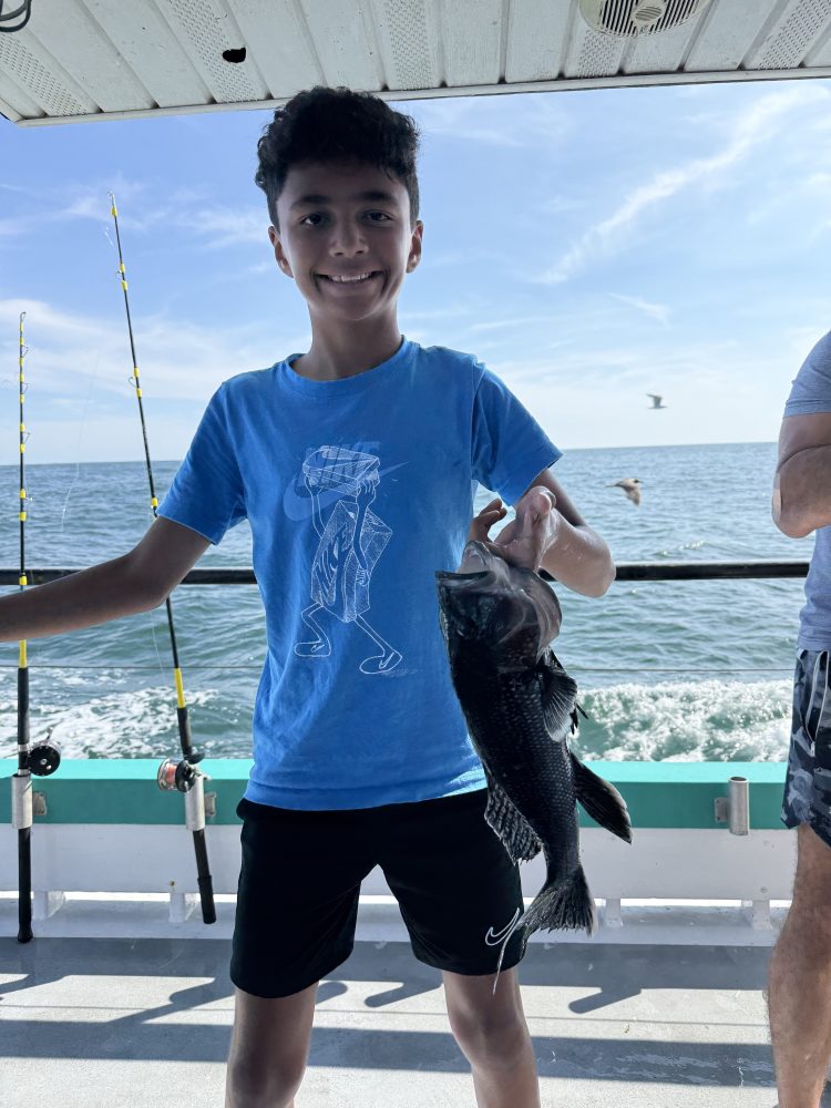 Boy on boat holding a fish, smiling, with ocean and fishing rods in background.