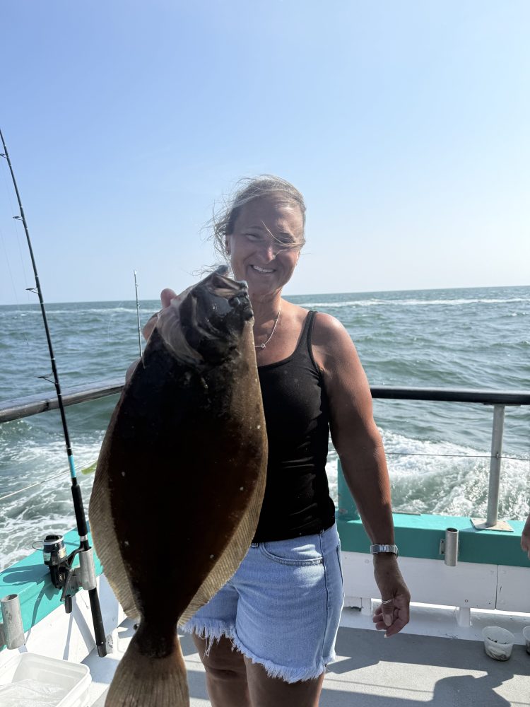 Smiling person on a boat holding a large fish against a backdrop of open water and clear sky.