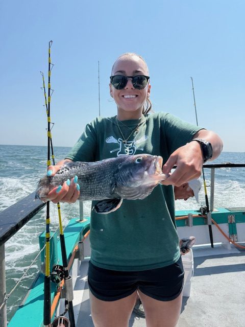 Woman on a boat holding a fish and smiling, with ocean and fishing rods in the background.
