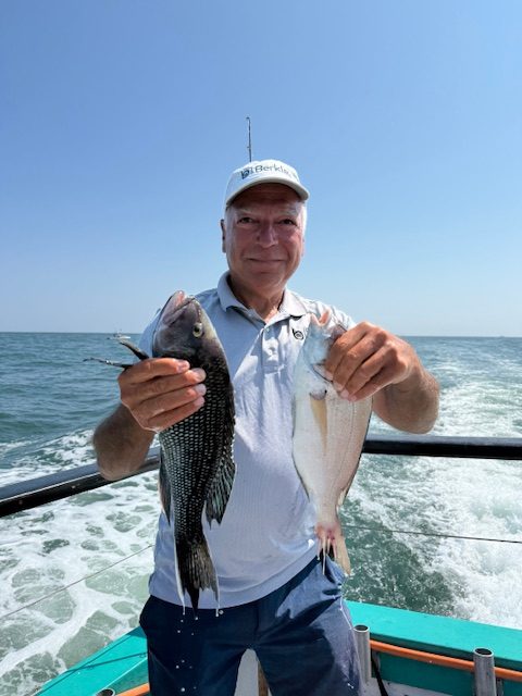 Man on a boat holding two fish against a sea backdrop under a clear blue sky.