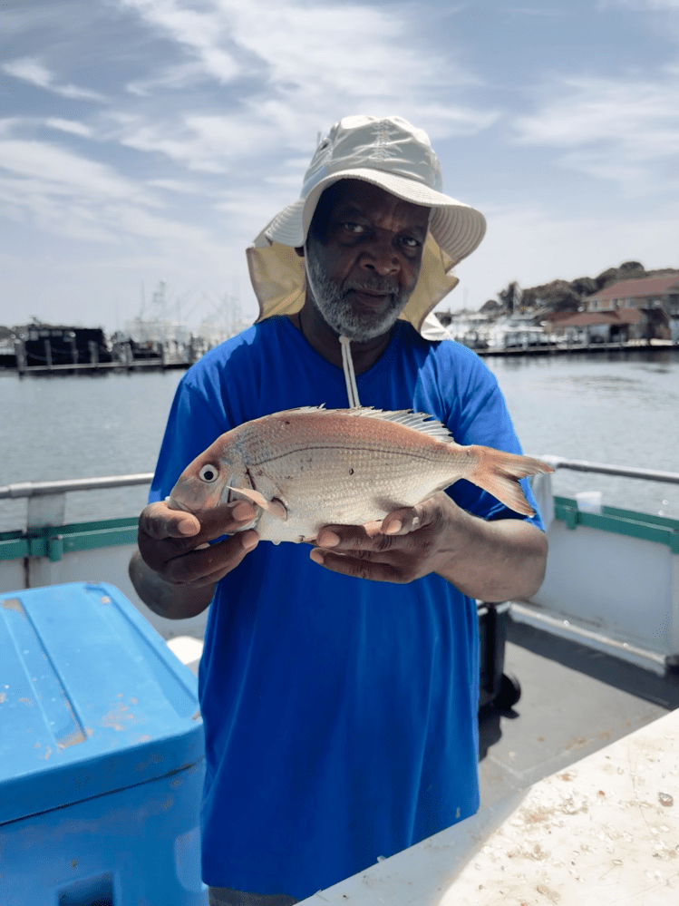 Man in blue shirt and hat holding a fish on a boat with water and buildings in the background.