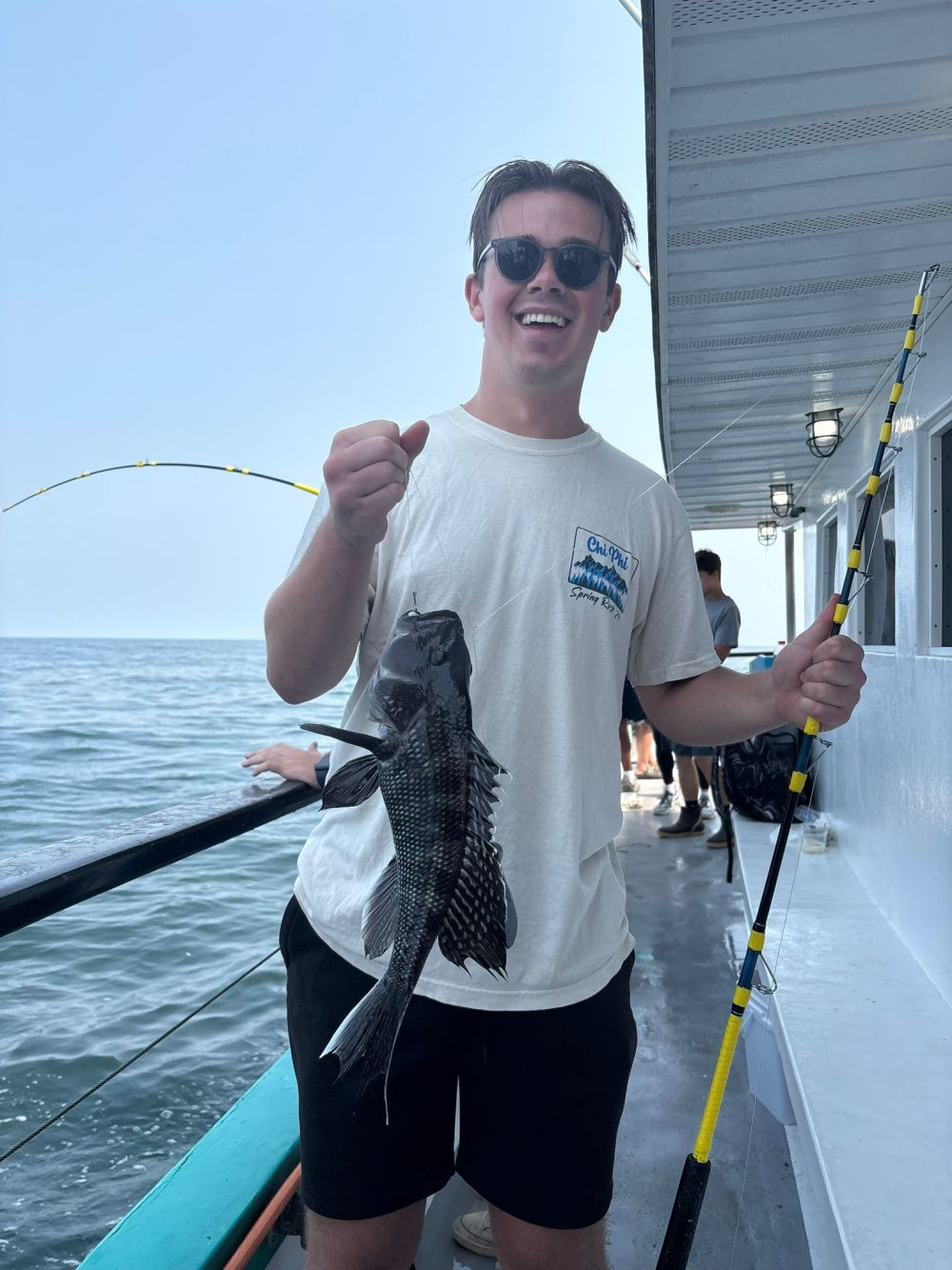 Person wearing sunglasses holding a fishing rod with a fish, standing on a boat.