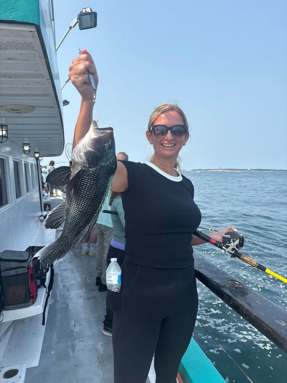 Woman on a boat holding a large fish with a fishing rod, smiling at the camera.
