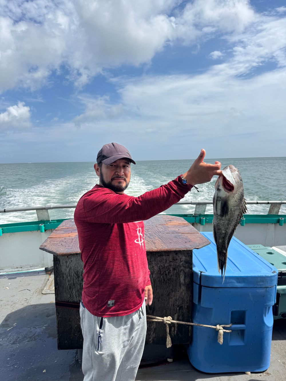 Man on a boat holding a fish, ocean and wake in the background.