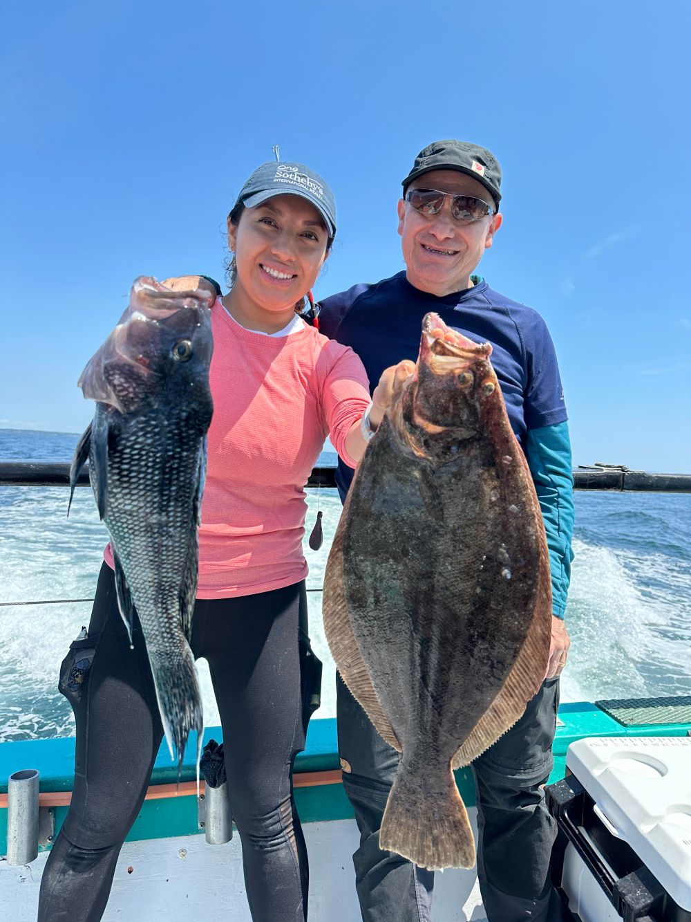 Two people smiling on a boat, holding a large fish each, under a clear blue sky.