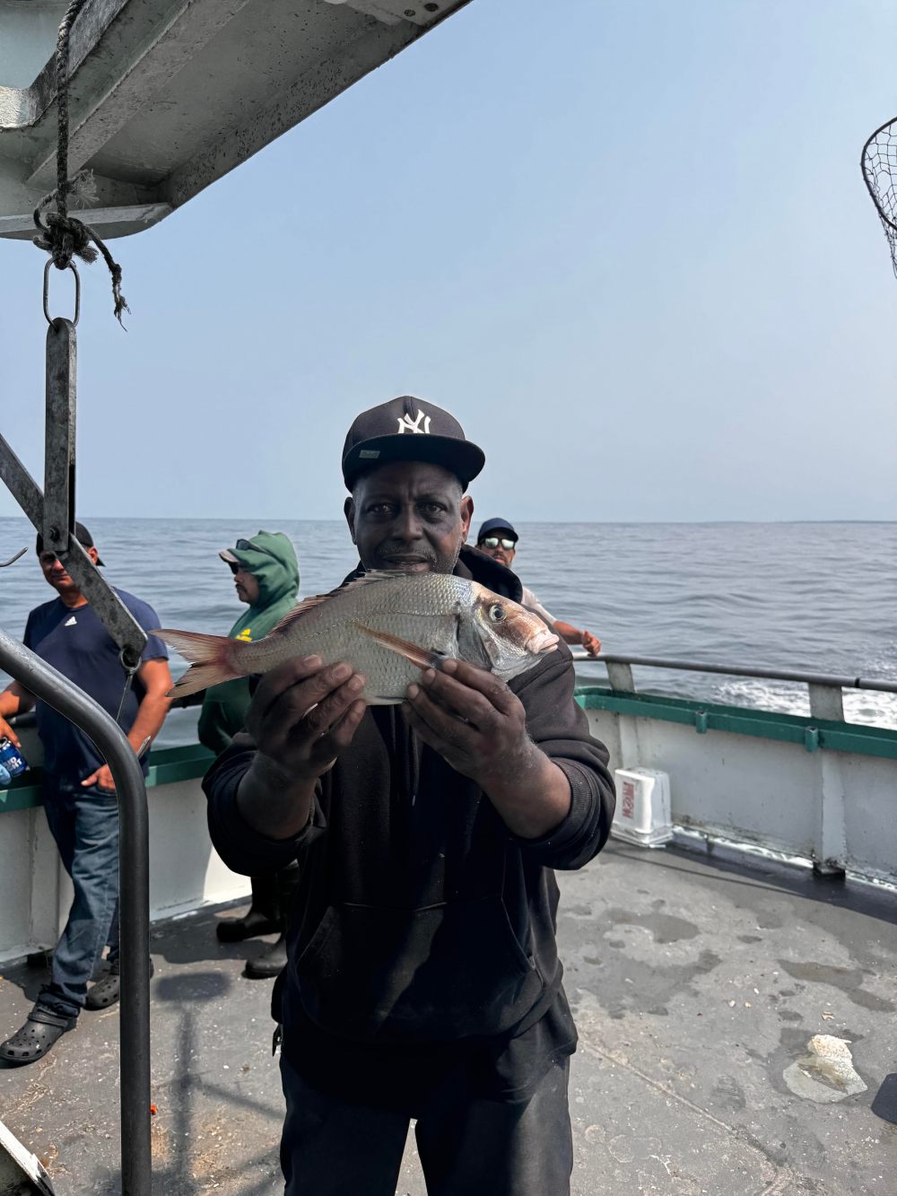 Person in black hoodie and cap holding a fish on a boat with ocean in background.