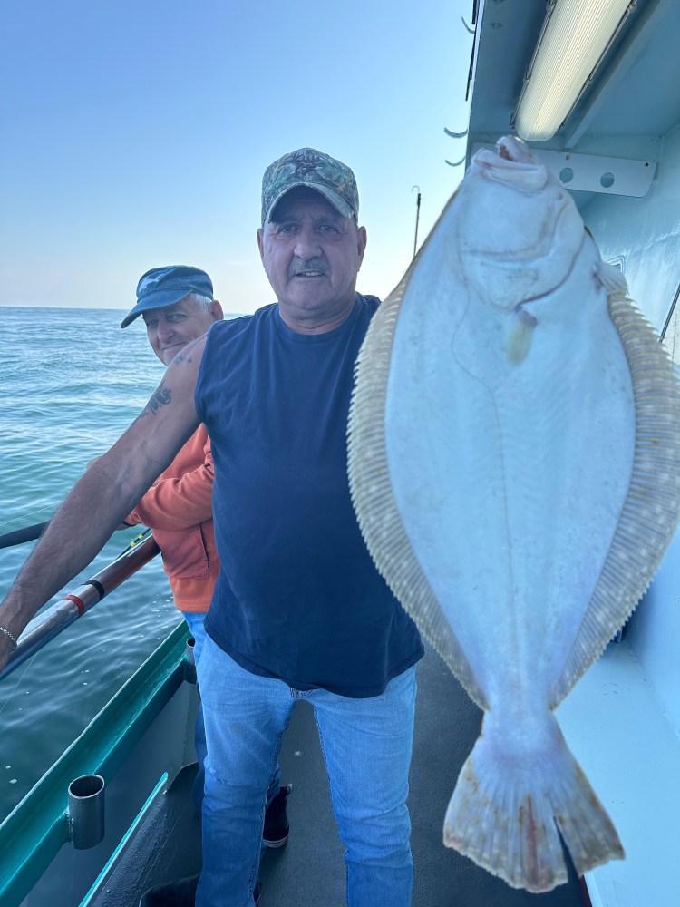 Two men on a boat, one holding a large flat fish, with ocean in the background.