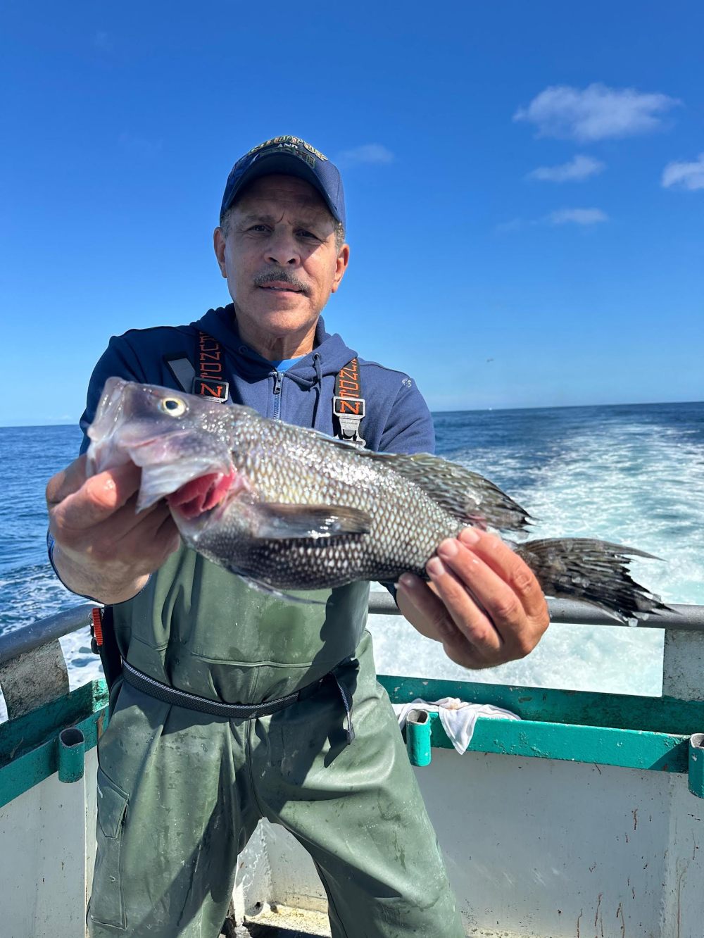 Man in blue cap holding a fish on a boat with ocean backdrop.