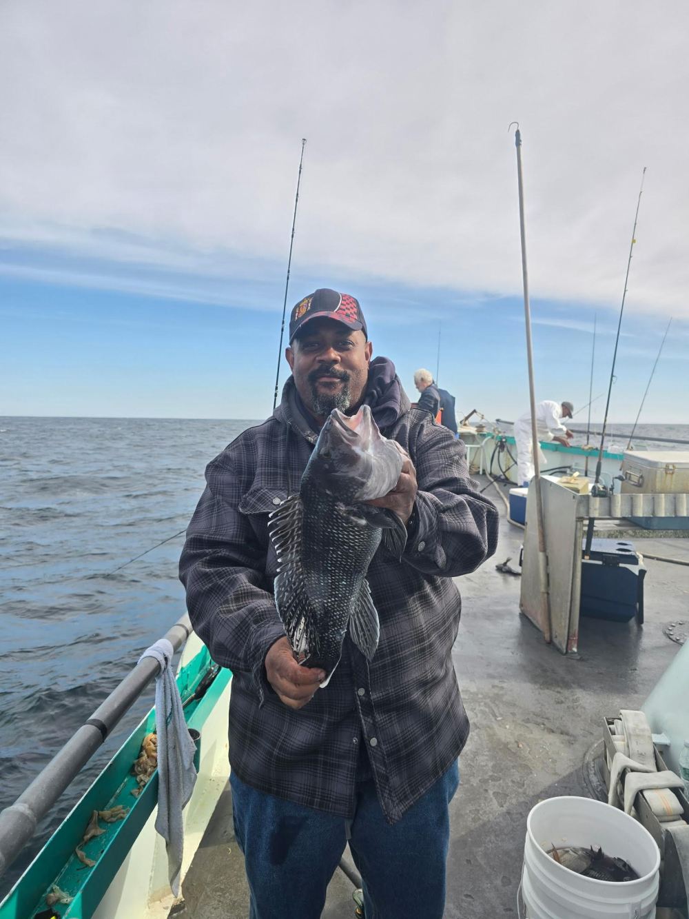 Man holding a fish on a boat with fishing poles and ocean in the background.