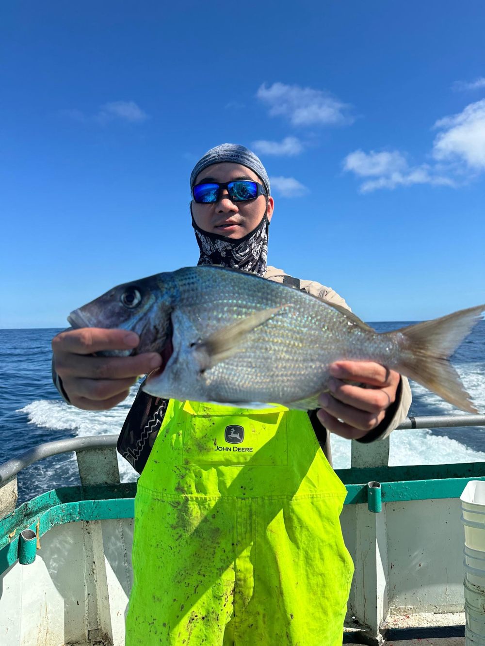 Person in bright yellow overalls holding a fish on a boat with ocean and blue sky background.