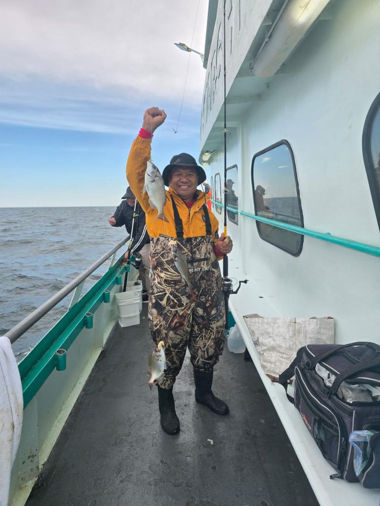 Person in camouflage gear holding two fish on a boat with ocean background.