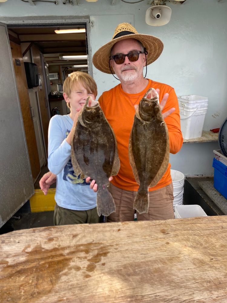 Two people holding large flounder fish on a boat, one wearing an orange shirt and straw hat.