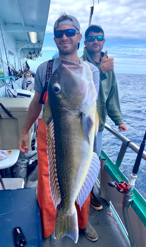 Two men on a boat, one holding a large fish, with the ocean in the background.