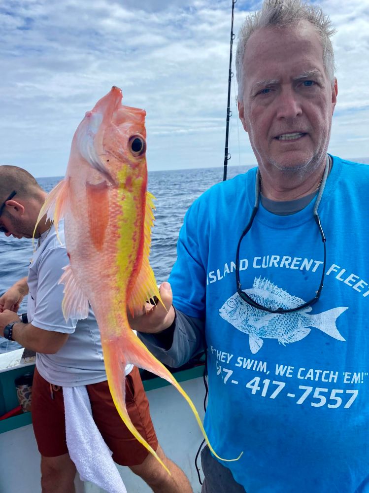 Man in a blue shirt holding a colorful fish on a boat with ocean in the background.