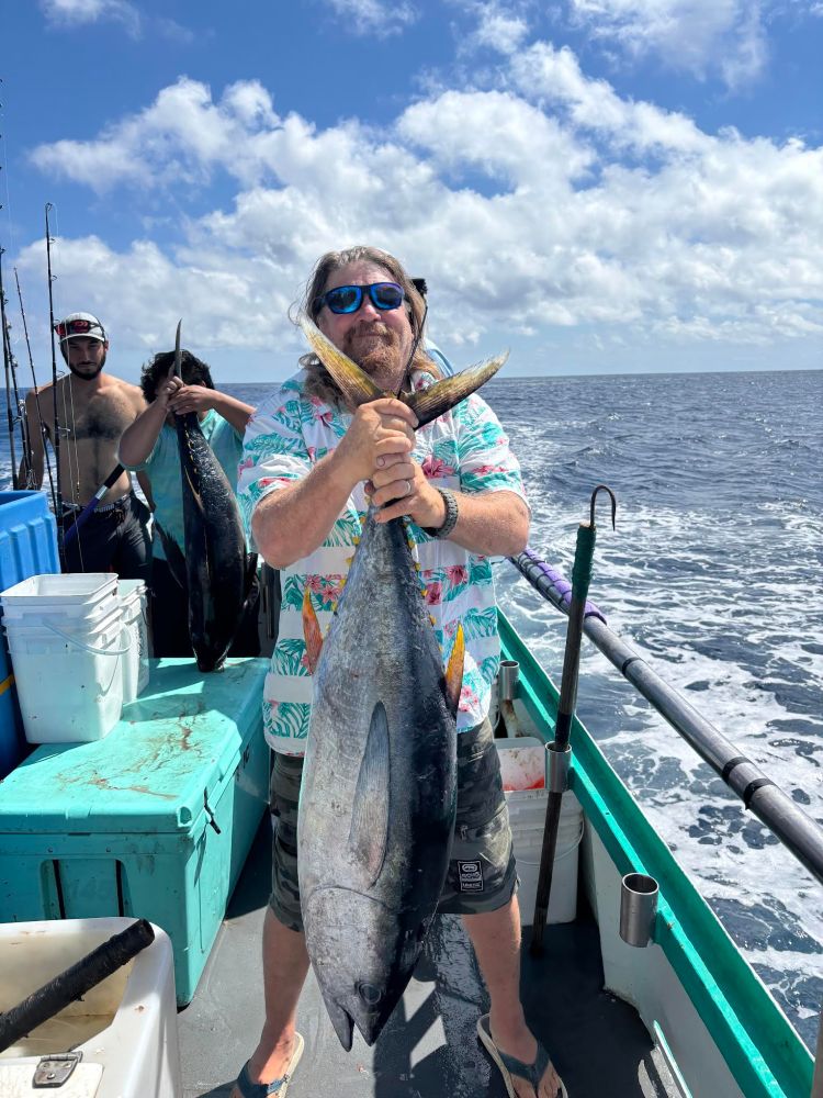 Man holding large tuna on a boat with others and fishing gear in the background under a blue sky.