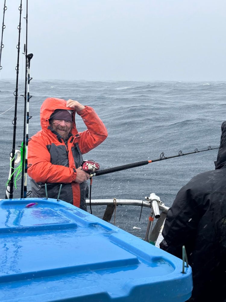 Person in orange raincoat fishing on a boat in rough seas, smiling despite the rain.