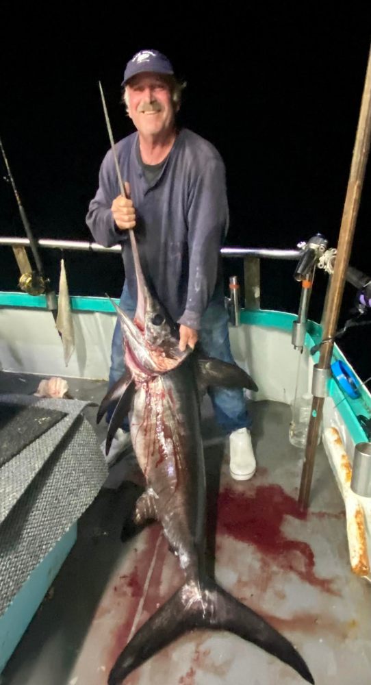 Man in hat holding a large swordfish on a boat at night.
