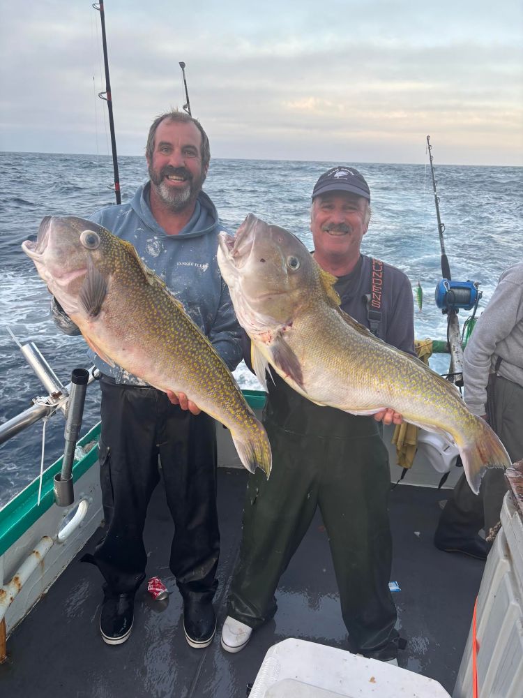 Two people on a boat holding large fish with ocean backdrop.