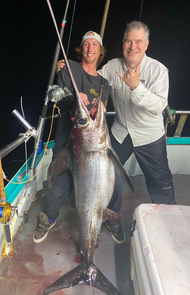 Two men on a boat holding a large swordfish they caught at night.