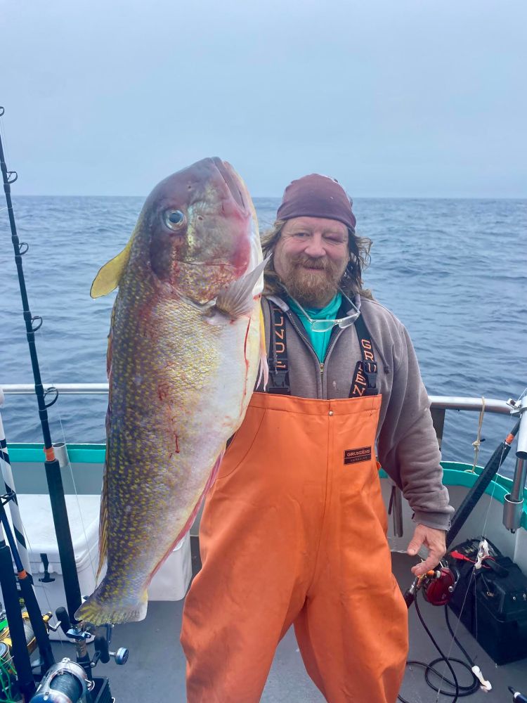 Person in orange overalls holding a large fish on a boat with the ocean in the background.