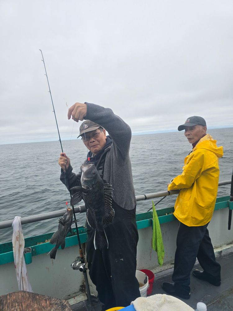 Two people on a boat; one holds a fishing rod with two caught fish, the other in a yellow raincoat watches.