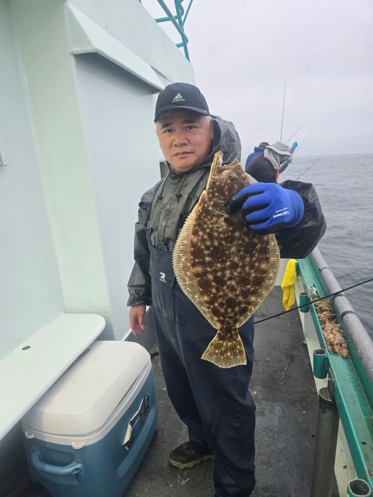 Person in rain gear holds a flatfish on a boat with a cooler and fishing rods visible.