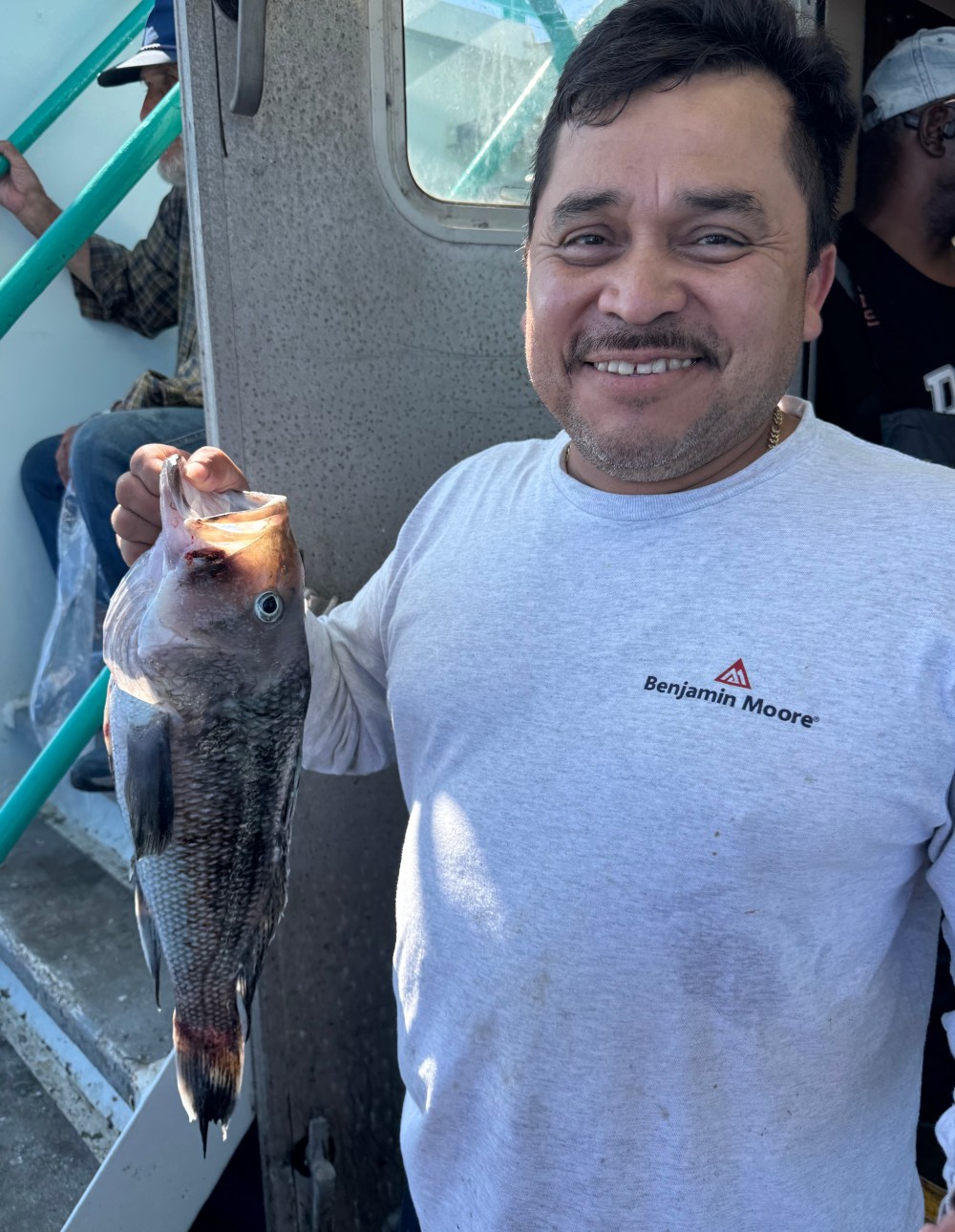 Smiling man holding a fish on a boat, wearing a gray shirt with 'Benjamin Moore' logo.