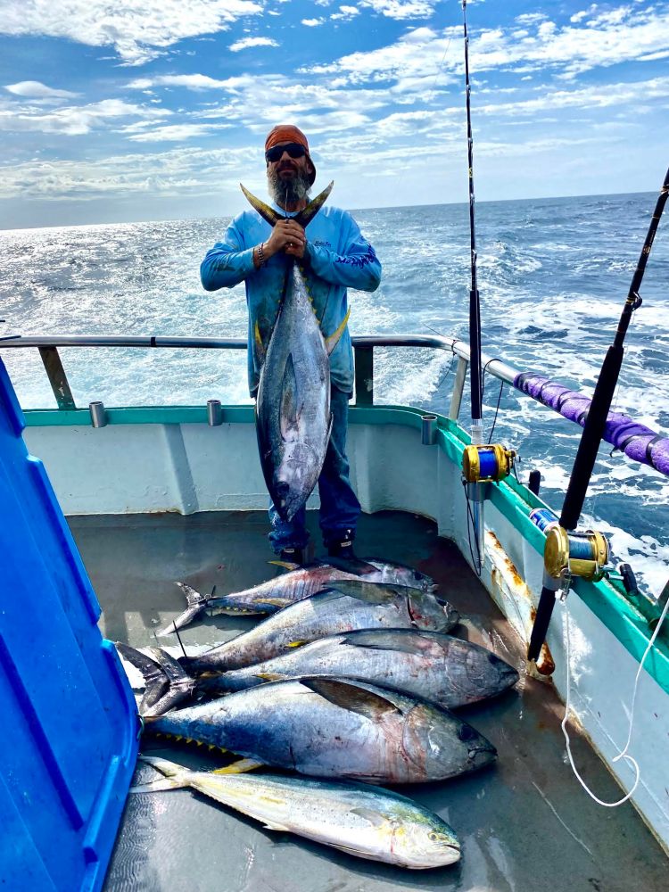 Person on a boat holding a large fish with several others on the deck, ocean and sky in the background.