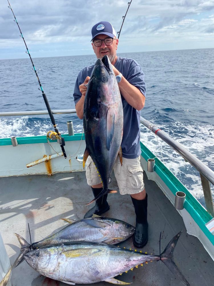 Person on a boat holding a large fish with two more fish on the deck, ocean in background.