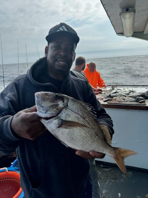 Man holding a fish on a boat, ocean in the background with people and fishing rods visible.