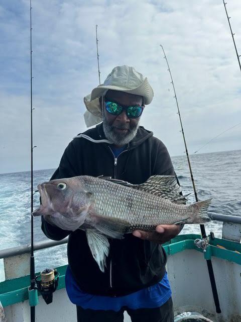 Person holding a fish on a boat, wearing a hat and sunglasses, with fishing rods in the background.