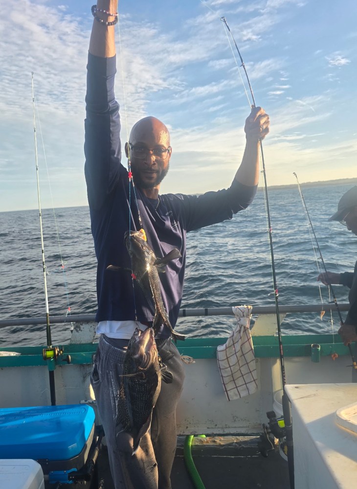 Person on a boat holding two fish with a fishing rod, ocean in the background.