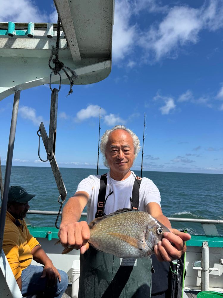 Man holding a fish on a boat with ocean in the background, under a sunny blue sky.
