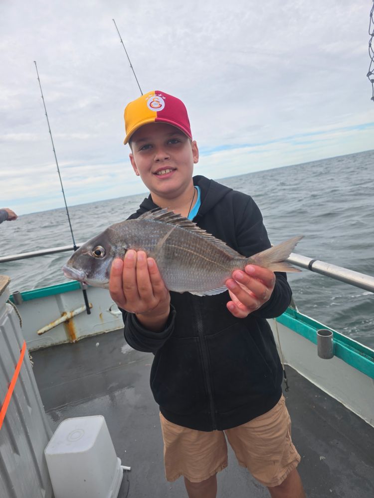 Child on a boat holding a fish, wearing a red and yellow cap and black jacket.