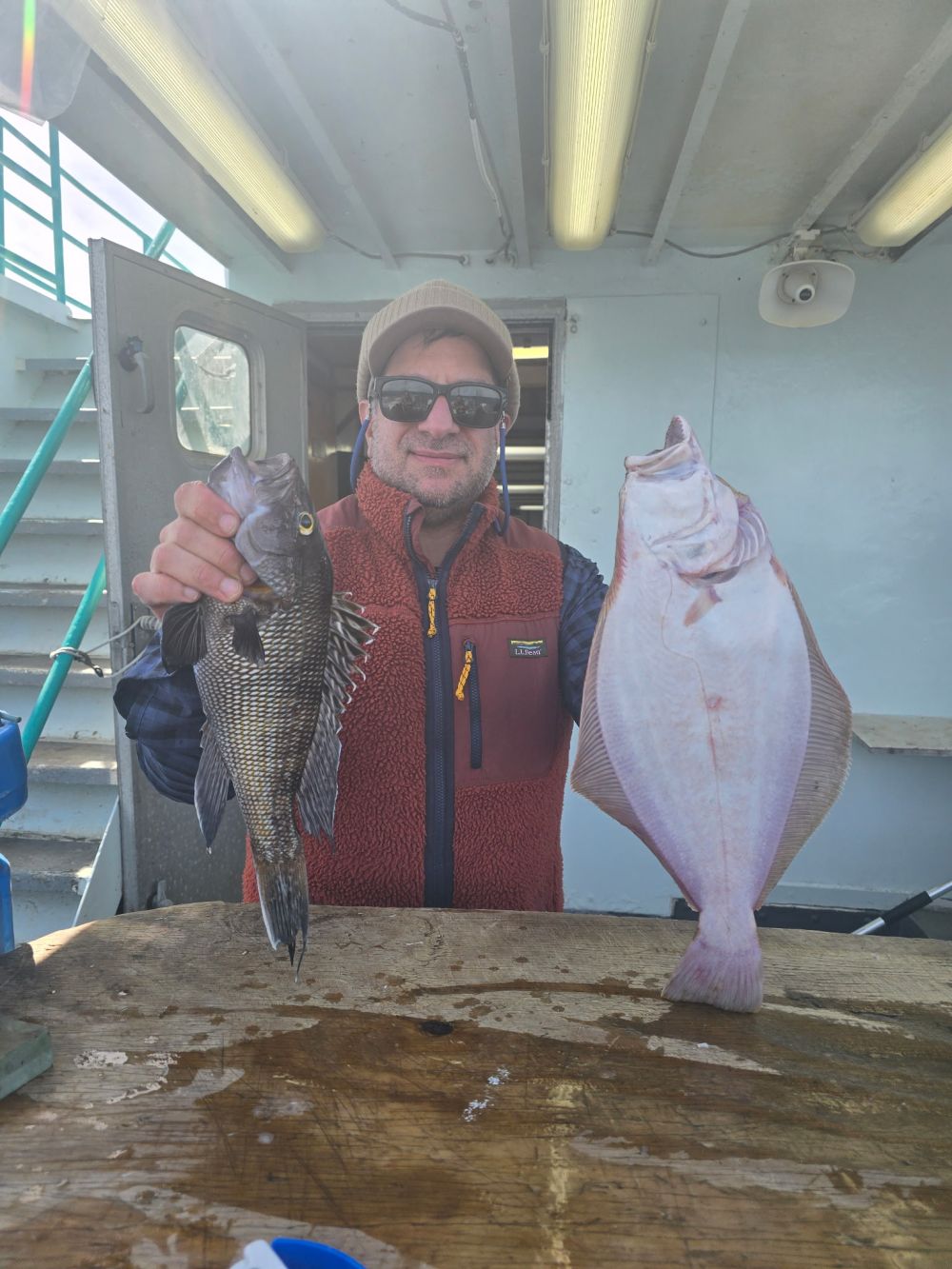 Person holding a sea bass in one hand and a flatfish in the other on a boat.