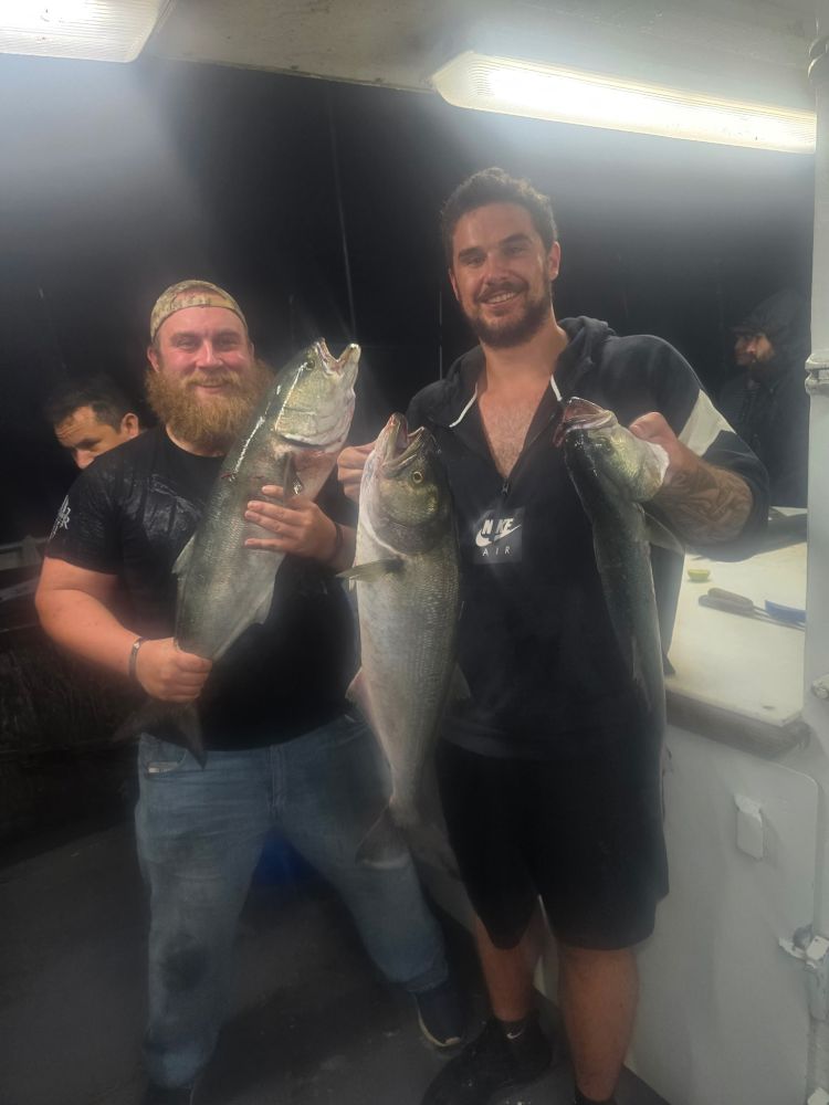 Two people holding large fish on a boat at night, smiling under bright lights.